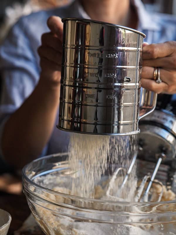 A person uses a metal flour sifter to sift flour into a clear mixing bowl below, with a mixer and dough visible in the bowl. The scene is warmly lit, and the persons face is out of focus.
